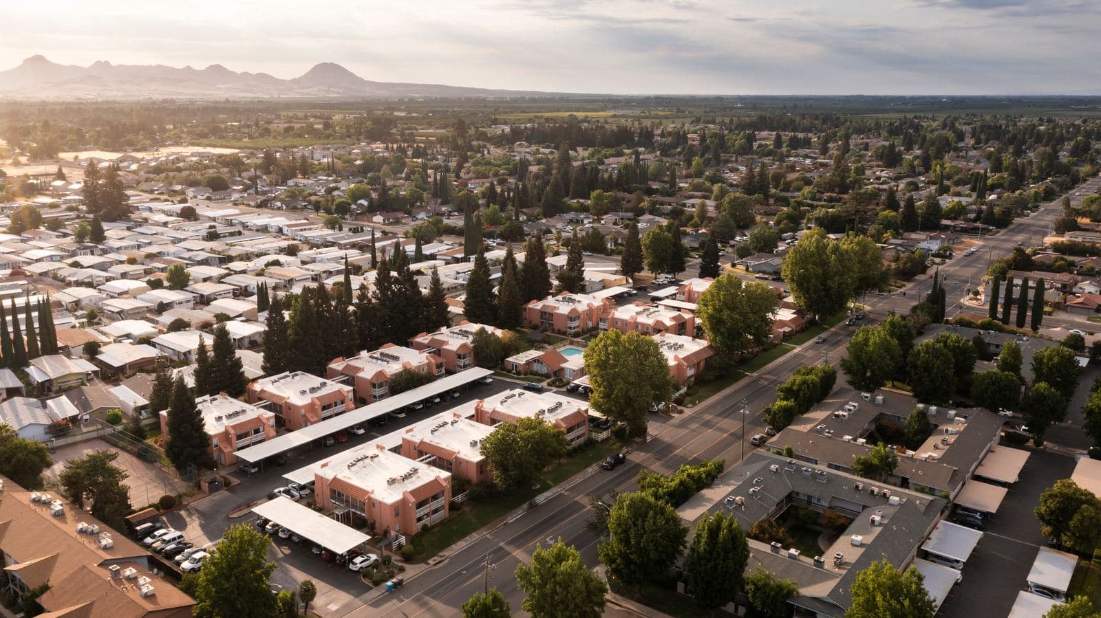 Aerial view over Yuba City, California at golden hour, with tree-lined residential streets and low apartment blocks below the Sutter Buttes on the horizon