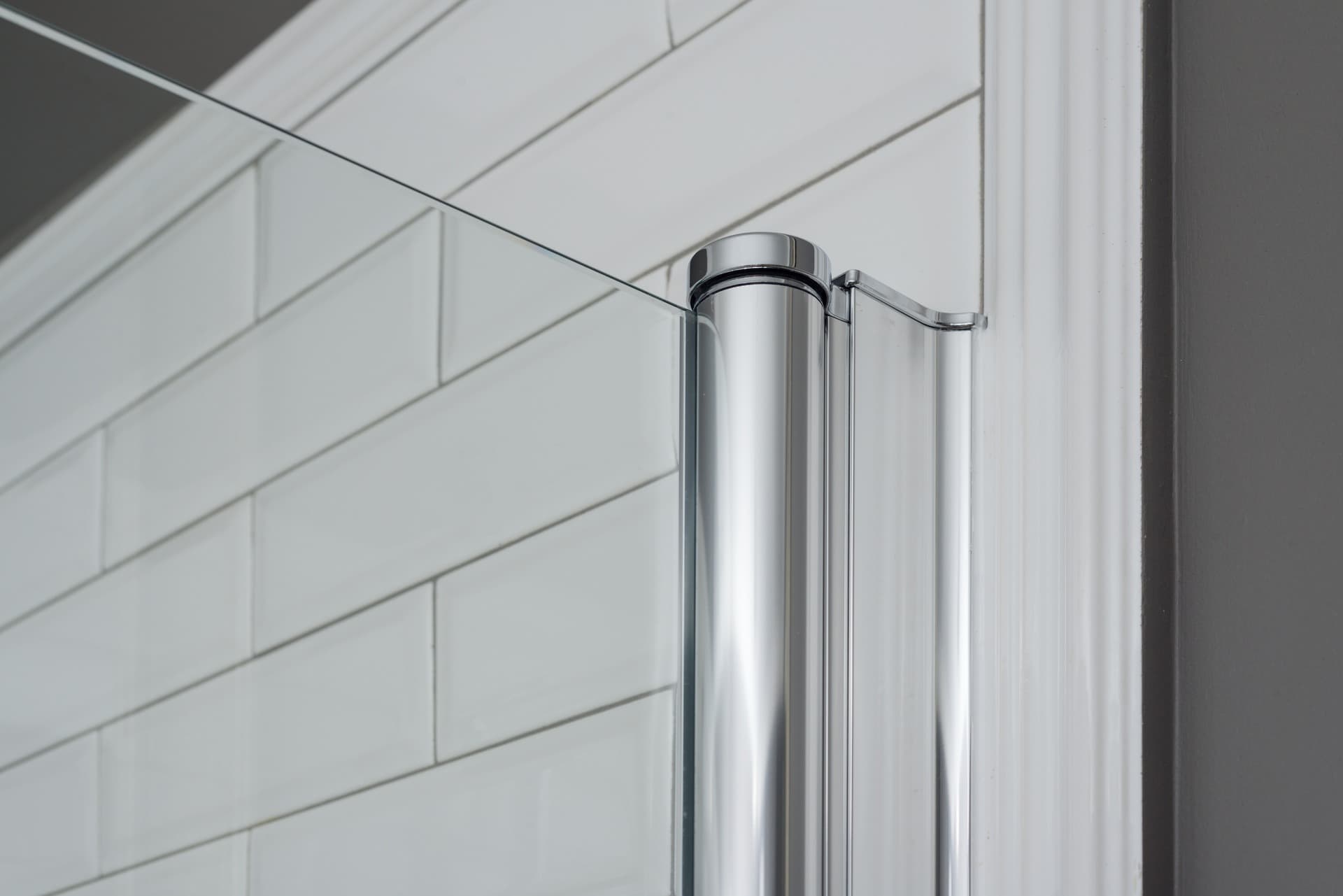 Close-up of a polished chrome shower door hinge holding a frameless glass panel against white tile