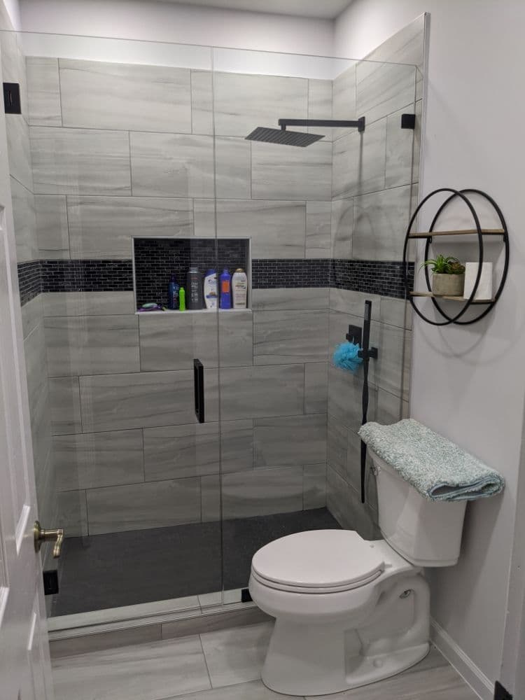 Sliding glass shower doors on a grey tile-lined walk-in shower with a black rain showerhead and a recessed toiletry niche